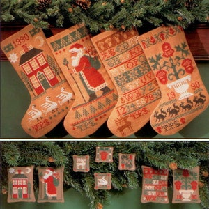 May include: Four beige Christmas stockings with cross-stitch designs, including a house, Santa Claus, and decorative patterns. The stockings are adorned with green, red, and white accents, and are hanging from a garland of pine needles and pine cones. Small matching ornaments are below.