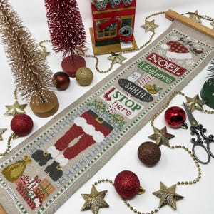 May include: A beige linen Christmas stocking with a Santa Claus design, the words "Stop here Santa", "I Believe", and "Noel" are embroidered on the stocking. The stocking is hanging on a wooden dowel rod.