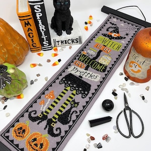 May include: A cross-stitch banner with a Halloween theme, featuring the text "Witch is in" and "Welcome my pretties." The design includes a witch's legs, pumpkins, and a black cat. The banner is displayed with Halloween decorations, such as a black cat figurine and candy corn.