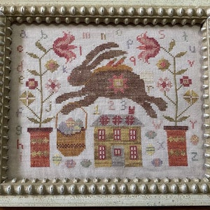 May include: Cross-stitch artwork in a white beaded frame. The design features a brown rabbit leaping over a house and basket of eggs. Floral elements and the alphabet surround the central image, with a color palette of browns, reds, and greens.