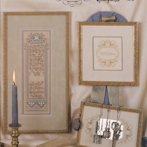 May include: An image featuring framed cross-stitch samplers and decorative items. The samplers include an alphabet and a welcome message, all in gold thread. A lit candle in a brass holder and antique scissors are also visible. The text "Cross 'N Patch" and "Elegant Alphabets I" are displayed.