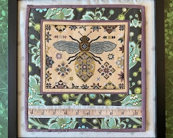 Puede incluir: Un bordado de punto de cruz enmarcado que presenta una abeja estilizada con un patrón geométrico. La abeja está rodeada de un diseño floral en tonos verdes, marrones y morados. El marco es negro y tiene una regla a lo largo del borde inferior.