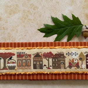 Könnte beinhalten: Ein Kreuzstich-Sampler, der eine Reihe von Häusern, Bäumen und einer Krähe zum Thema Herbst zeigt, vor einem beigen Hintergrund. Der Sampler ist mit orangefarbenem Stoff und einem Fransenrand eingefasst. Ein Eichenblatt und eine Eichel befinden sich im Hintergrund.