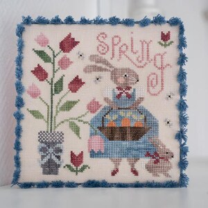 May include: Cross-stitch artwork featuring a bunny holding a basket of carrots, with the word "SPRING" stitched above. The design includes tulips and a blue fringe border. The artwork is square and measures approximately 8 inches by 8 inches.