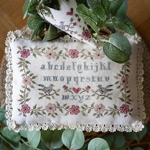 Könnte beinhalten: Ein weißes Kissen mit einer Spitzenborte zeigt ein Kreuzstich-Design mit einem floralen Rand mit rosa Blüten und grünen Blättern. Die Mitte des Kissens zeigt ein Kreuzstich-Alphabet mit zwei Vögeln und einem Herzen.