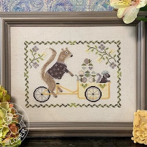 May include: A cross-stitch embroidery of a squirrel riding a bicycle with a basket full of colorful eggs. The squirrel is wearing a brown jacket and has a bushy tail. The bicycle has a yellow basket and wheels. The background is a green and white floral design.