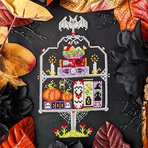 May include: A cross-stitch design featuring a white shelf filled with Halloween-themed items. The shelf holds pumpkins, books, candles, and a bat. The design is set against a black background with autumn leaves.