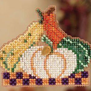 Op de afbeelding: Een kruissteekdecoratie met een pompoen, maïs en een wortel. De pompoen is wit met oranje accenten, de maïs is geel en groen en de wortel is oranje. De basis heeft een geblokt patroon.