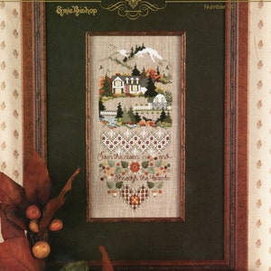 May include: Framed cross-stitch artwork featuring a landscape scene with houses, trees, and mountains. The artwork includes the text "Over the river and Through the broodo" and is set in a dark green mat within a brown wooden frame. Decorative leaves and berries surround the frame.