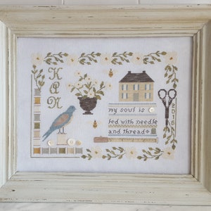 Puede incluir: Obra de arte de punto de cruz en un marco de madera blanco envejecido. El diseño presenta un pájaro azul, una casa, flores, tijeras y el texto "my soul is fed with needle and thread". La obra incluye carretes de hilo y botones.