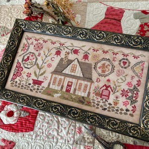 May include: Framed cross-stitch sampler featuring a house, flowers, and birds in a detailed, folk art style. The sampler is in a dark, ornate frame. The color palette includes beige, red, green, and brown. The sampler is displayed on a quilted background.