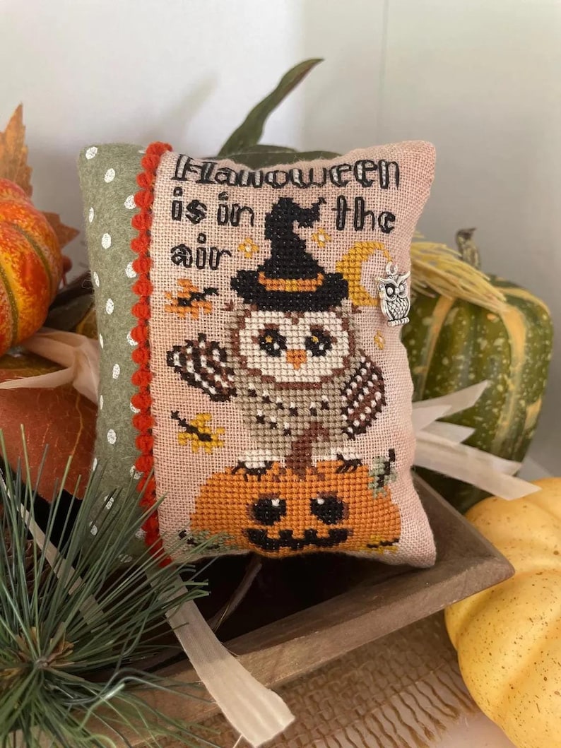 May include: A pink and green cushion with a cross-stitch design featuring an owl wearing a witch's hat and sitting on a pumpkin. The text "Halloween is in the air" is stitched on the cushion.