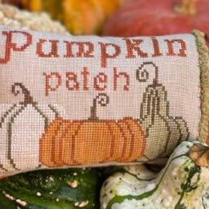 May include: A rectangular, cross-stitched pillow with the words "Pumpkin Patch" in orange thread. Three pumpkins in white, orange, and green are stitched below the text. The pillow is surrounded by gourds and pumpkins.