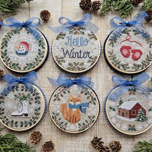 Peut inclure: Six décorations circulaires en tambour à broder avec des motifs au point de croix sur tissu beige clair. Les motifs incluent un bonhomme de neige, le texte "Hello Winter", des moufles rouges, un patin à glace blanc, un renard orange et un chalet rouge.