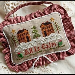 May include: A rectangular fabric pincushion featuring a cross-stitched winter scene with two brown houses, green pine trees, and snow, with the text All Is Calm. The cushion is bordered by a red and white gingham ruffled trim and a hanging loop.