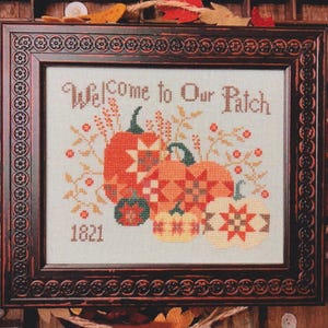 May include: Cross-stitch artwork in a dark brown frame. The artwork features pumpkins in shades of orange, yellow, and green, with the text "Welcome to Our Patch" and the number "1821". The pumpkins have star and geometric patterns.