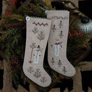 Könnte beinhalten: Zwei beige Weihnachtssocken mit gestickten Schneemännern und Bäumen. Die Strümpfe haben einen gewellten Rand und sind mit Knöpfen verziert. Die Strümpfe sind von Grün und Tannenzapfen umgeben und schaffen eine festliche Atmosphäre.