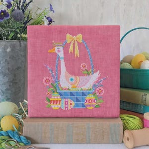 May include: A pink cross-stitch piece features a goose in a blue basket with Easter eggs and flowers. The goose wears a crown and a yellow bow adorns the basket. The artwork is displayed on a stack of books, with Easter eggs and flowers in the background.