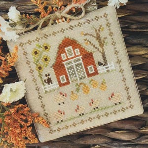 Könnte beinhalten: Kreuzstich-Stickmuster mit einem roten Bauernhaus, Sonnenblumen, Kürbissen und Schafen. Das Design ist mit einem dekorativen Rand versehen und umfasst einen Baum und einen kleinen Zaun. Die Farbpalette umfasst Orange-, Grün- und Beigetöne.