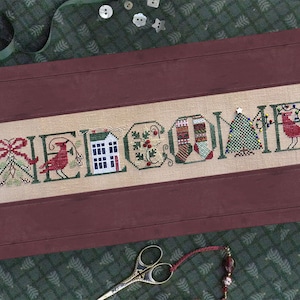 Peut inclure: Broderie au point de croix encadrée dans un cadre rouge foncé. Le mot « WELCOME » est brodé en fil vert, chaque lettre représentant un motif de vacances, notamment un cardinal, une maison et un sapin de Noël. Le fond est en tissu beige clair.