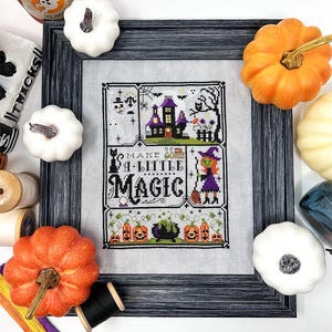 May include: A framed cross-stitch sampler on light gray fabric featuring a haunted house, a witch with a broom, a cauldron, and pumpkins. The text reads "MAKE A LITTLE MAGIC" in black lettering. The piece is surrounded by decorative pumpkins.