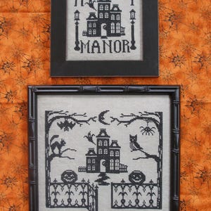 May include: Two framed cross-stitch embroideries on light fabric. The top piece features a house with the text "MIDNIGHT MANOR". The bottom piece depicts a house, a crescent moon, a bat, a spider, a cat, an owl, and two jack-o'-lanterns.