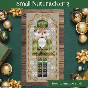 May include: Cross-stitch pattern of a nutcracker soldier in green, red, and white. The text "Small Nutcracker 3" is at the top. The stitch count is 36w x 79h. The design is surrounded by gold and white snowflake accents. Christmas ornaments and bows surround the design.