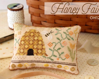 May include: A cross-stitched cushion with a beehive and bee design. The cushion has a beige background with a detailed pattern of a yellow and brown beehive, a white and black bee, and floral accents. The text "Honey Faire" is displayed.