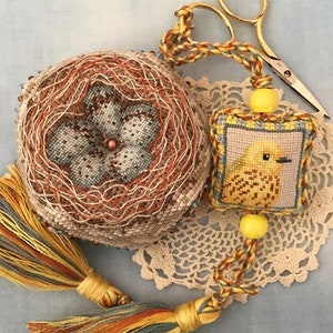 May include: A yellow bird cross stitch pattern with a brown and beige nest pincushion and a yellow tassel. The bird is stitched on a small square piece of fabric with a light blue border.
