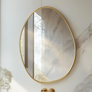 Puede incluir: Un espejo ovalado con marco dorado cuelga sobre un lavabo con diseño de mármol. El lavabo tiene el texto "AC MIRROR". La escena del baño incluye un grifo, dispensadores de jabón y toallas enrolladas, lo que sugiere un diseño moderno y minimalista.