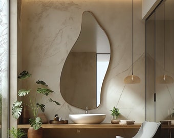 May include: A contemporary bathroom featuring a white sink and a large, organically shaped mirror. The mirror is framed in a thin, gold-coloured metal. The bathroom has a light wood floor and a white wall with a subtle marble pattern. There are plants in the room, and a wicker pendant light hangs from the ceiling.