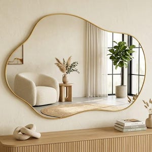 Op de afbeelding: Een onregelmatig gevormde wandspiegel met een dunne, goudkleurige metalen rand. Het reflecterende oppervlak toont een kamerinterieur met een crèmekleurige fauteuil, een kleine houten bijzettafel met droogbloemen en een hoge kamerplant.