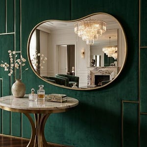 May include: An elegant interior scene featuring a large, uniquely shaped gold-framed mirror reflecting a chandelier and other furnishings. A marble-topped table with a gold base holds decorative items. The wall is a deep green with gold accents, and there are two armchairs.