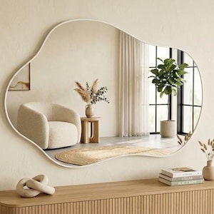 May include: A large wall mirror with an irregular, organic, cloud-like shape and a thin white frame. The mirror reflects a room containing a cream-colored armchair, a small wooden side table with dried plants, and a tall green potted plant.