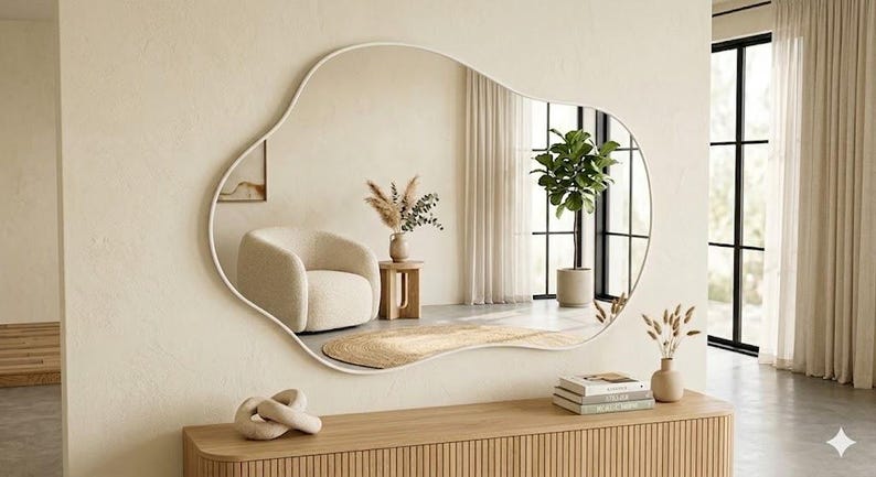 May include: A large wall mirror with an irregular, organic, cloud-like shape and a thin white frame. The mirror reflects a room containing a cream-colored armchair, a small wooden side table with dried plants, and a tall green potted plant.