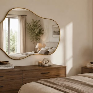 May include: An asymmetrical wall mirror with a thin gold frame featuring an irregular, organic shape. The mirror is mounted on a wall above a wooden dresser, reflecting a bedroom interior with a window, curtains, and a potted plant.