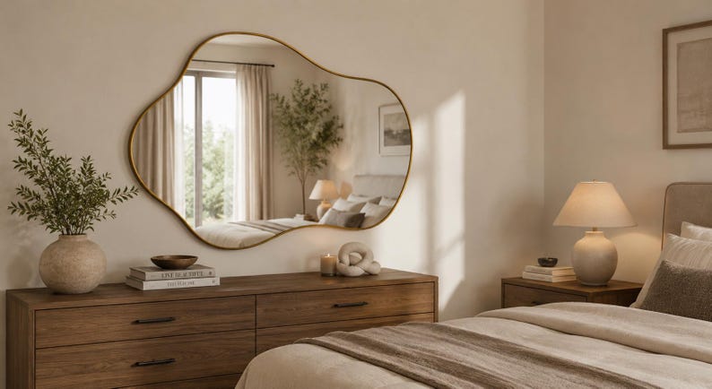 May include: An asymmetrical wall mirror with a thin gold frame featuring an irregular, organic shape. The mirror is mounted on a wall above a wooden dresser, reflecting a bedroom interior with a window, curtains, and a potted plant.
