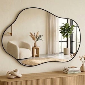 May include: A large wall mirror with an irregular, organic, blob-like shape and a thin black frame. The mirror is mounted on a light beige wall above a wooden console table, reflecting a room with a cream-colored armchair and a potted plant.