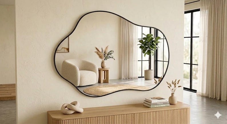May include: A large wall mirror with an irregular, organic, blob-like shape and a thin black frame. The mirror is mounted on a light beige wall above a wooden console table, reflecting a room with a cream-colored armchair and a potted plant.