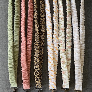 May include: Six fabric lanyards with gathered, scrunchie-style designs. The lanyards feature various patterns and colors: solid sage green, dusty pink with white dots, black and tan leopard print, white with orange carrots, white with small pink flowers, and solid white.