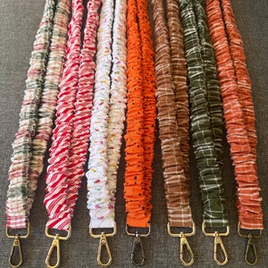 May include: Seven fabric lanyards with metal swivel clasps, featuring various patterns including green and red plaid, red and white stripes, white with colorful holiday lights, orange with black dots, brown plaid, green plaid, and orange plaid designs.