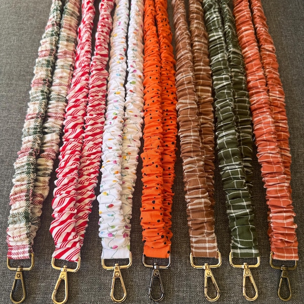 May include: Seven fabric lanyards with metal swivel clasps, featuring various patterns including green and red plaid, red and white stripes, white with colorful holiday lights, orange with black dots, brown plaid, green plaid, and orange plaid designs.