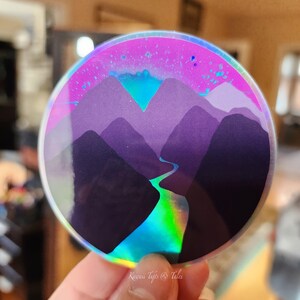 May include: A round iridescent sticker with a design of purple mountains and a blue river flowing through them. The background is a pink and blue gradient with white stars.