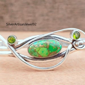 Op de afbeelding: Een zilveren manchetarmband met een ovale turkooissteen in het midden, met groene en bruine tinten. Twee kleinere groene stenen zijn aan weerszijden gezet. De armband heeft een uniek, vloeiend ontwerp.