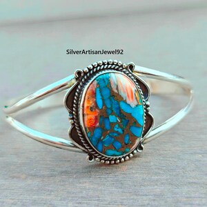 Könnte beinhalten: Ein silbernes Armreif-Armband mit einem ovalen, türkisfarbenen und orangefarbenen Stein im Zentrum. Der Stein ist in einem dekorativen Rahmen mit einem Perlenrand gefasst. Das Armband hat ein schlichtes, elegantes Design.