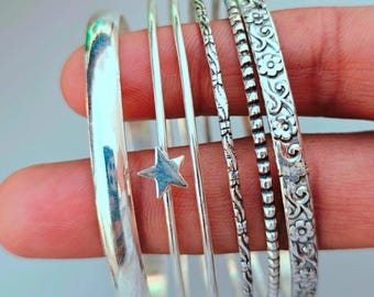 May include: A collection of silver bangles, including a thick, polished band, a star-shaped charm, and several thinner bands with textured and floral designs. The bangles are displayed on a hand.