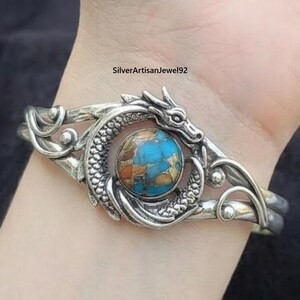 Könnte beinhalten: Ein silberfarbenes Armband mit einem Drachendesign. Der Drache umschließt einen runden, türkis- und kupferfarbenen Stein. Das Armband wird am Handgelenk getragen und zeigt die filigranen Details des Schmucks.