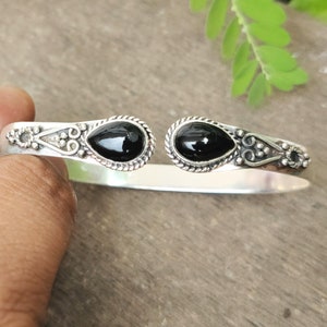 Könnte beinhalten: Ein silbernes Armreif-Armband mit zwei schwarzen Edelstein-Akzenten. Das Armband hat ein zartes, verziertes Design.
