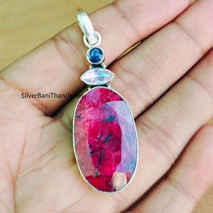 May include: A silver pendant featuring a large oval-shaped red ruby gemstone, surrounded by a smaller oval-shaped clear gemstone and a round blue gemstone.