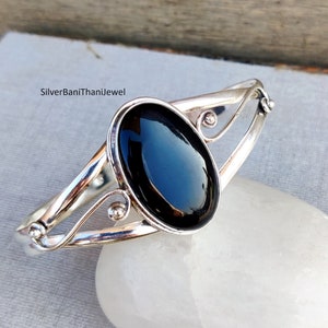 May include: A silver-toned bangle bracelet with an oval, glossy black stone at the center. The bracelet features a decorative, openwork design with curved lines and small, round accents. The text "SilverBaniThaniJewel" is visible.